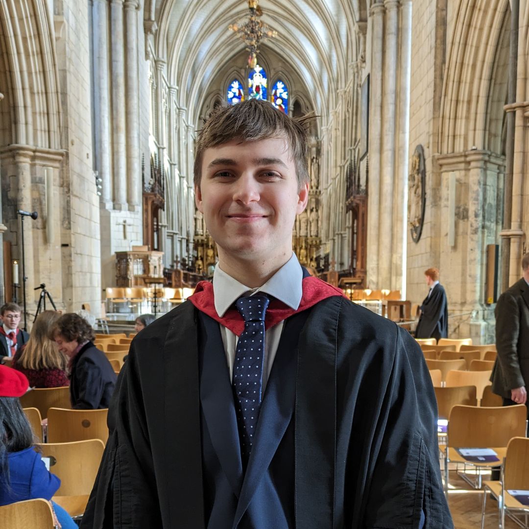 An image of Joel Colyer, wearing graduation robes over a suit and tie, in the nave of a cathedral with chairs visible on either side, and a stained glass window visible behind.