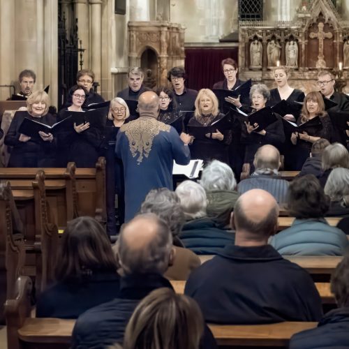 A choir of mixed genders wearing all black, standing in two rows in a church, facing the conductor, with the audience visible on pews in the foreground.